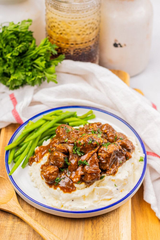 A plate of mashed potatoes topped with beef stew and fresh herbs, served with green beans, on a wooden tray with a spoon and a bunch of parsley nearby.