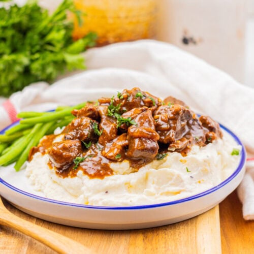 A plate of mashed potatoes topped with beef stew and garnished with green beans, served on a wooden board with a wooden spoon and fresh parsley in the background.