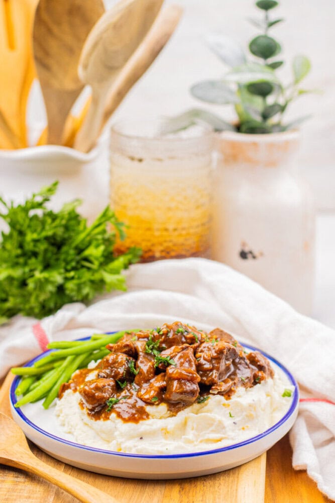 A plate of mashed potatoes topped with beef stew, garnished with green beans and chopped herbs, sits on a wooden board with kitchen utensils and plants in the background.