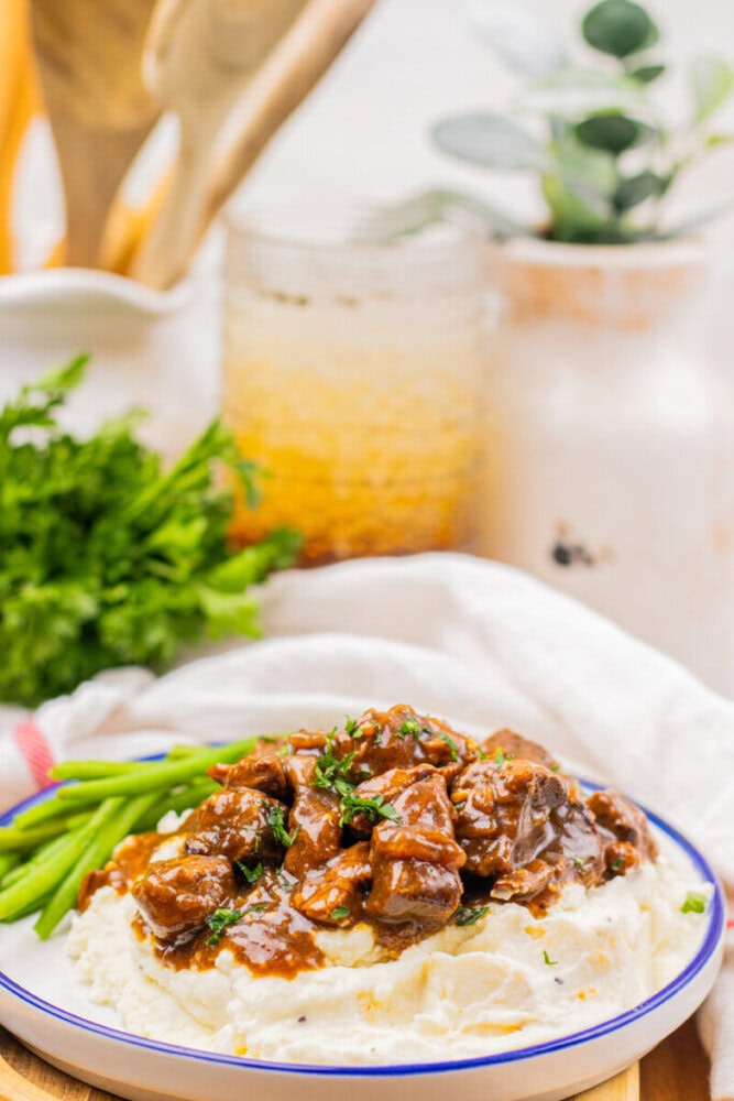 A plate of mashed potatoes topped with beef stew and garnished with herbs, served with green beans. Fresh parsley and kitchen items are visible in the background.