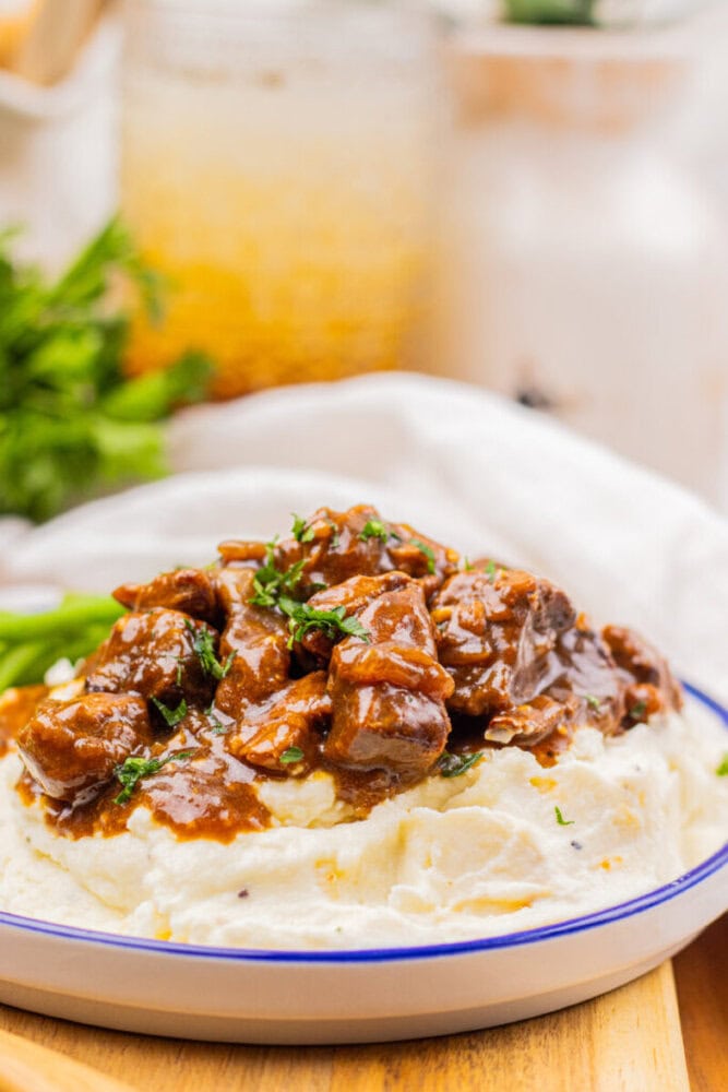 A plate of mashed potatoes topped with beef stew and garnished with chopped herbs, served on a white plate with a blue rim.