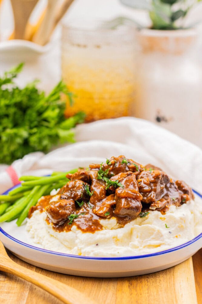 A plate of mashed potatoes topped with beef stew and garnished with herbs, served with green beans on the side.