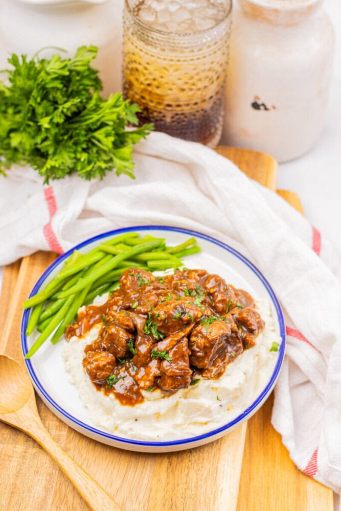 A plate of mashed potatoes topped with beef stew, served with green beans, on a wooden board with a bunch of parsley and a glass in the background.