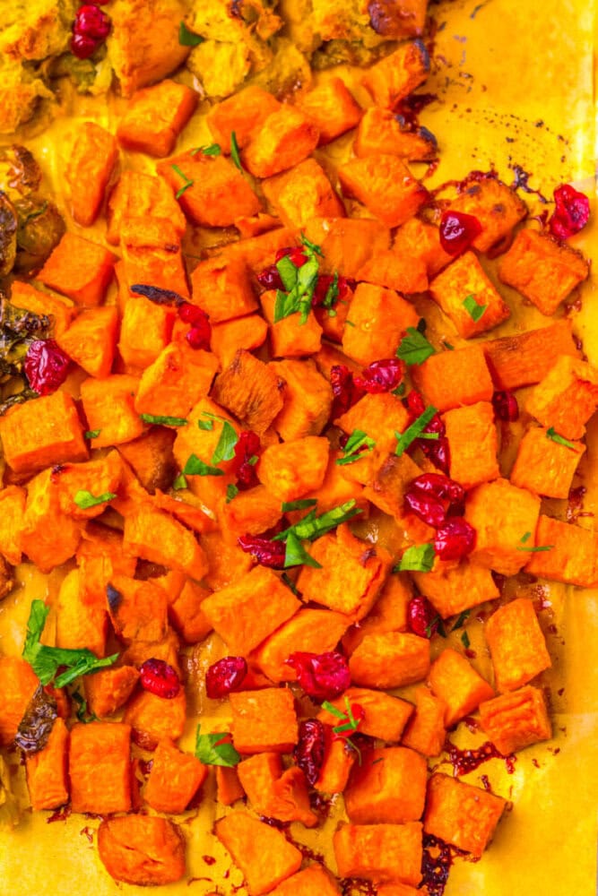 Roasted sweet potato cubes garnished with chopped parsley and cranberries on a yellow baking sheet.
