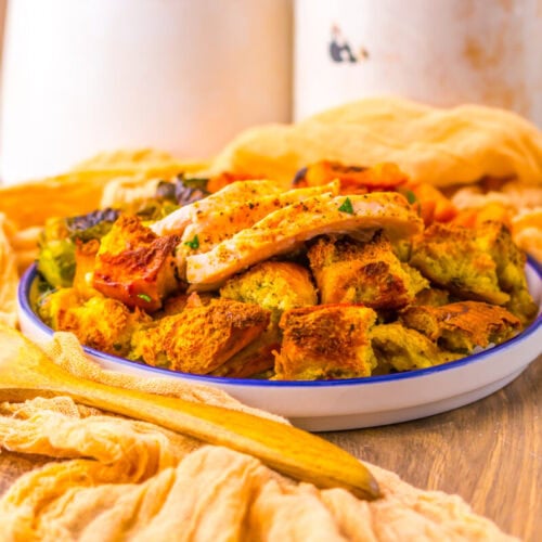 A plate of roasted vegetables and bread cubes topped with sliced grilled chicken, with a wooden spoon and yellow cloth on a wooden surface.