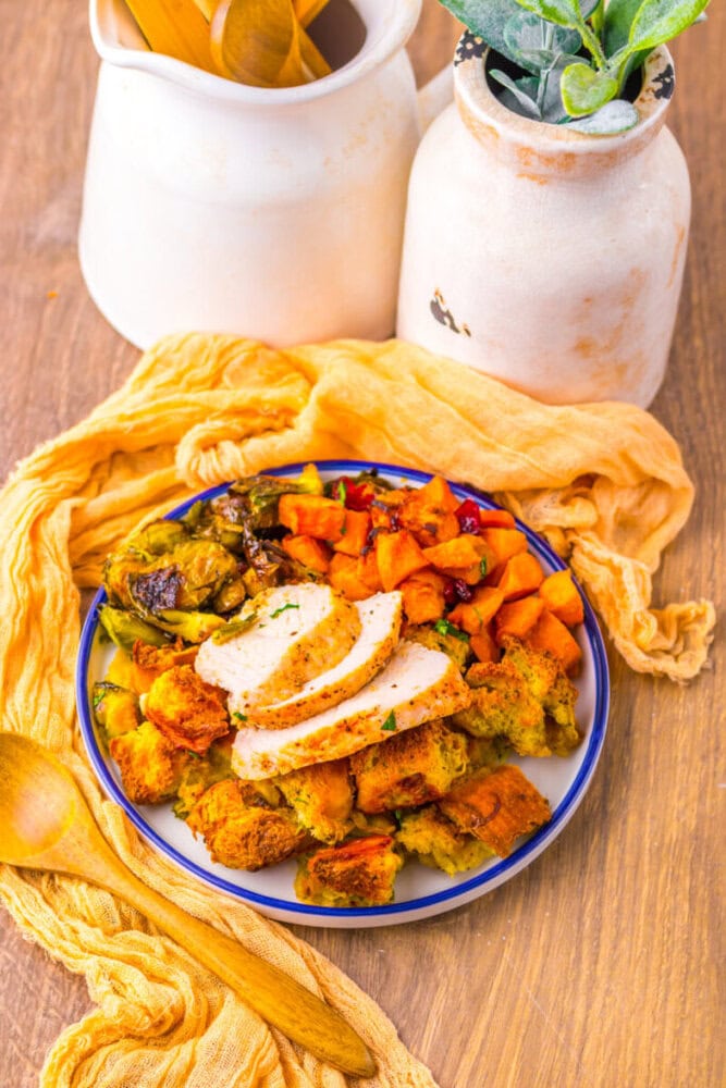 A plate with sliced roasted turkey, stuffing, roasted vegetables, and sweet potatoes, placed on a wooden table with ceramic jars and a yellow cloth.
