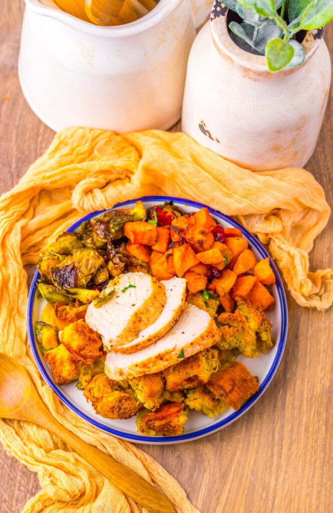 A plate with sliced turkey breast, roasted sweet potatoes, stuffing, and Brussels sprouts on a wooden table with a yellow cloth and ceramic jars.