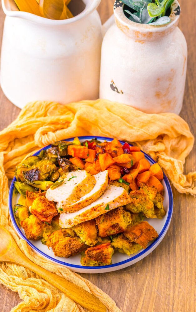 A plate with sliced turkey, roasted sweet potatoes, Brussels sprouts, and stuffing sits on a wooden table with a yellow cloth and white ceramic jars in the background.