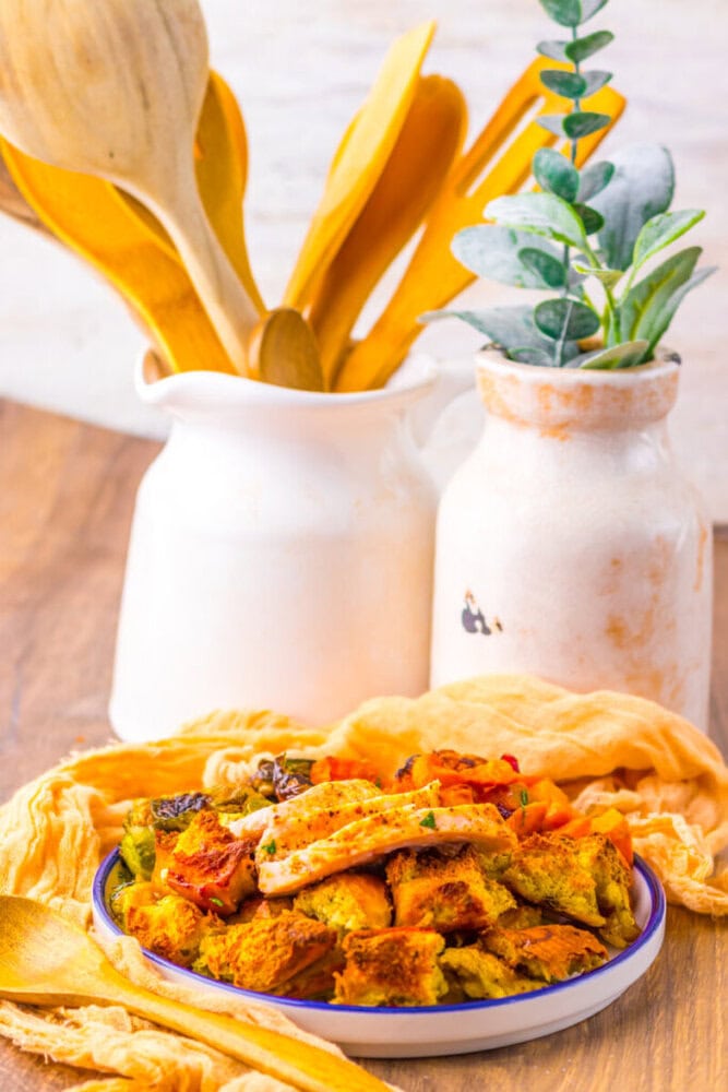 A plate of seasoned, roasted chicken pieces sits on a yellow cloth, with white ceramic jugs holding wooden utensils and a leafy plant in the background.