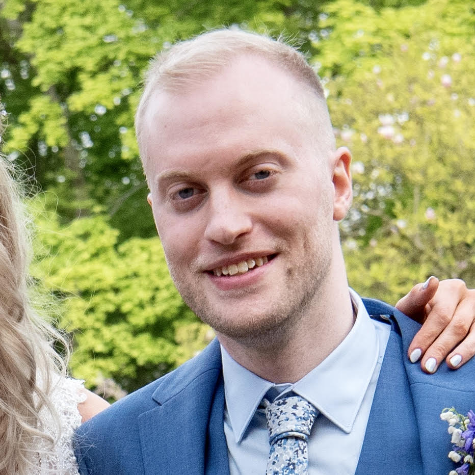 A man with short blond hair and a trimmed beard smiles while wearing a blue suit, white shirt, and patterned tie outdoors.