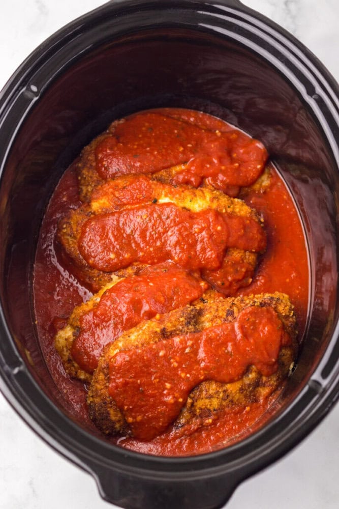 Breaded chicken breasts topped with tomato sauce inside a slow cooker, ready to be cooked.