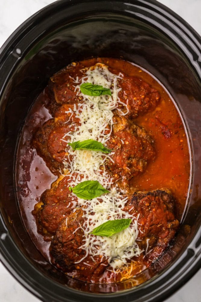 Four pieces of chicken covered in tomato sauce and melted cheese, topped with basil leaves, in a slow cooker.