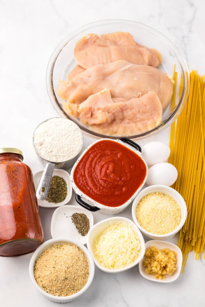 Raw chicken breasts, dry spaghetti, jarred tomato sauce, flour, eggs, shredded cheese, grated cheese, breadcrumbs, minced garlic, and dried herbs arranged on a white surface.