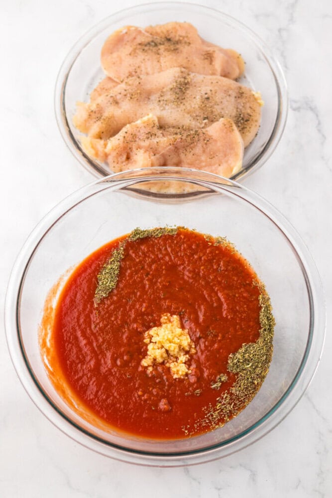 Two glass bowls on a marble surface; one contains raw seasoned chicken breasts, the other holds tomato sauce with herbs and minced garlic.