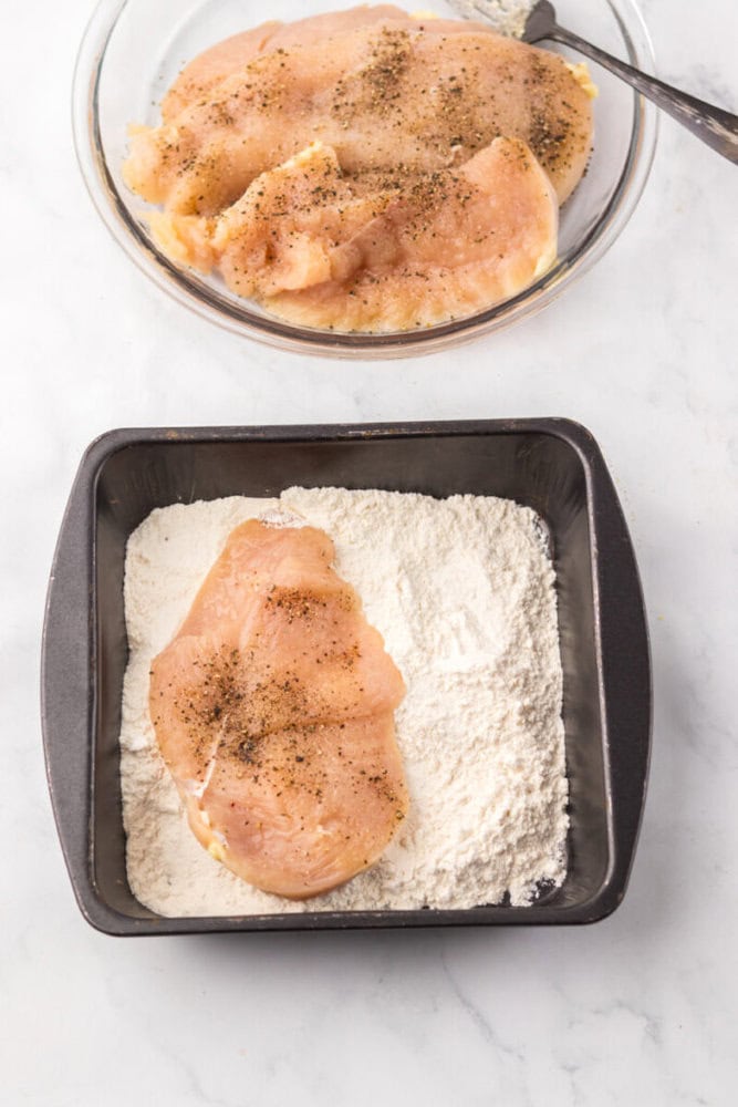 Raw, seasoned chicken breasts being coated in flour; one piece is in a square pan with flour, others are in a glass bowl above on a white surface.
