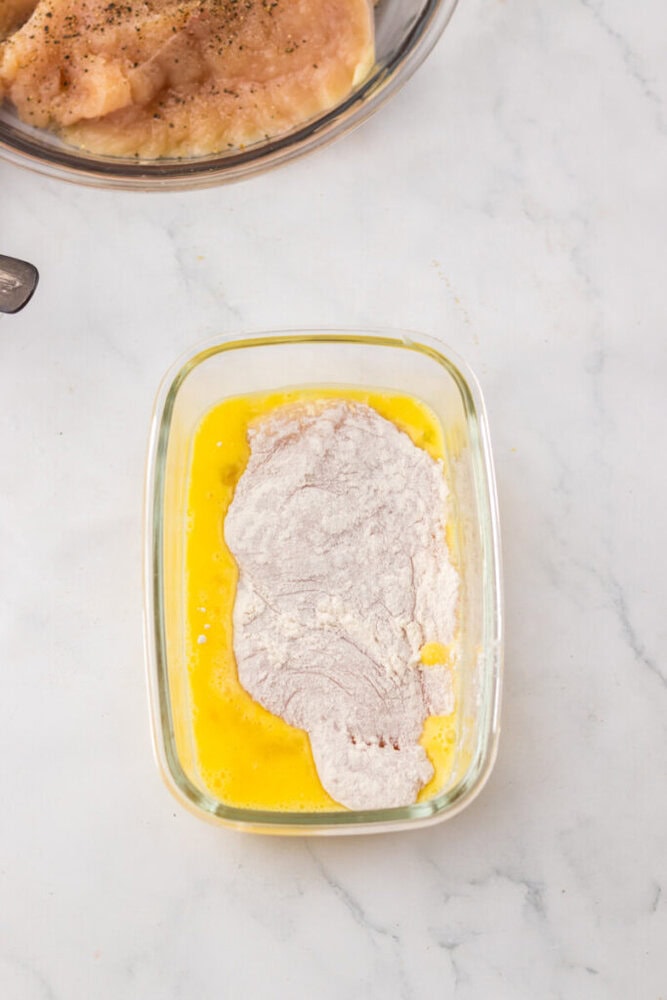 A piece of floured chicken breast is being dipped in a glass container of beaten eggs, with more raw chicken visible in a bowl above.