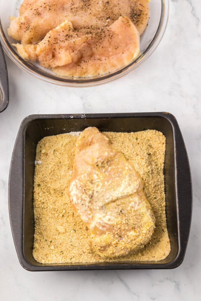 Raw chicken breast being coated in seasoned breadcrumbs in a square baking pan, with more chicken breasts in a glass bowl above.