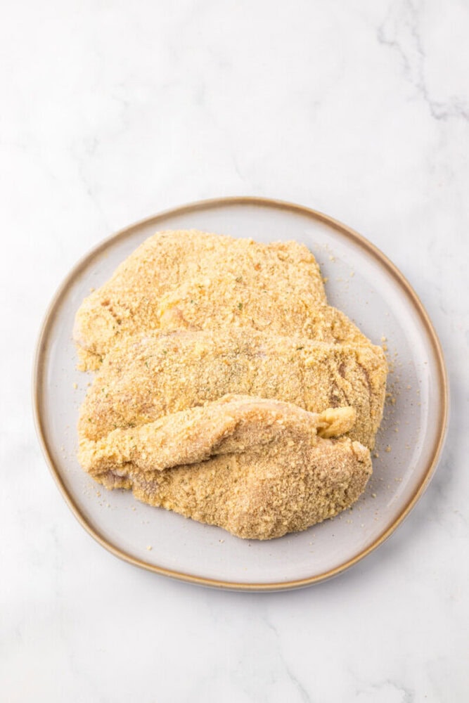 Three breaded pieces of raw chicken arranged on a round plate, set on a white marble surface.