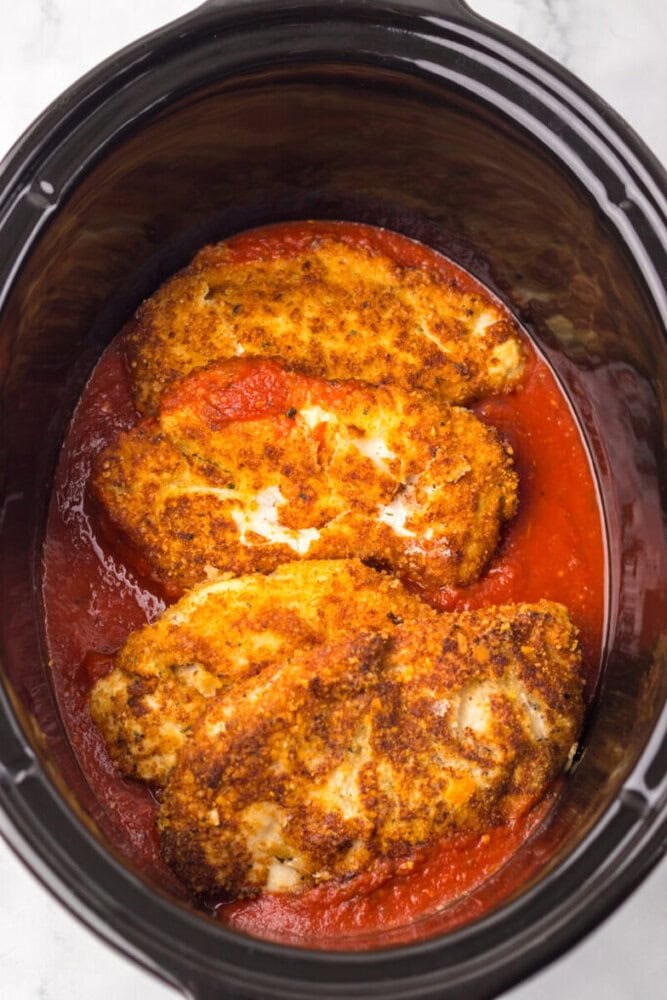 Three breaded chicken breasts in tomato sauce inside a black slow cooker.
