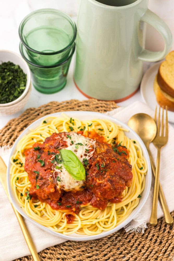A plate of spaghetti topped with tomato sauce and cheese sits on a woven placemat, with a gold fork and spoon, glassware, green pitcher, and a bowl of herbs nearby.