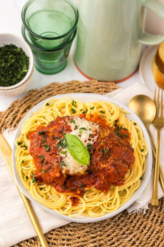 A plate of spaghetti topped with tomato sauce, a piece of chicken, grated cheese, and a basil leaf, served with a gold fork and knife on a woven placemat.