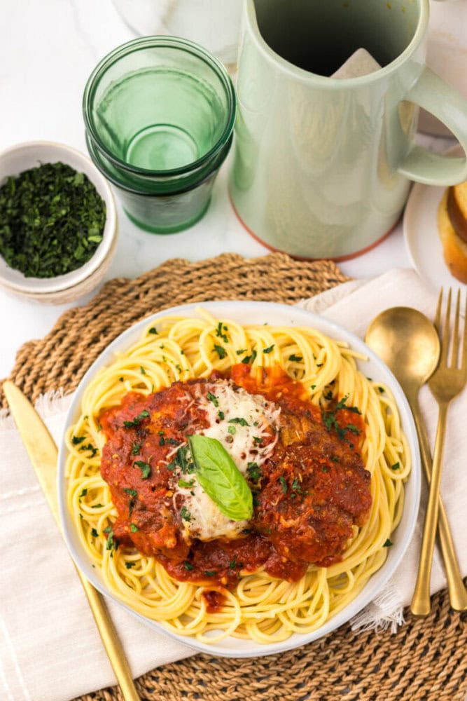 A plate of spaghetti topped with marinara sauce, melted cheese, and basil, set on a woven placemat with a fork, knife, napkin, green cup, and a bowl of chopped herbs nearby.