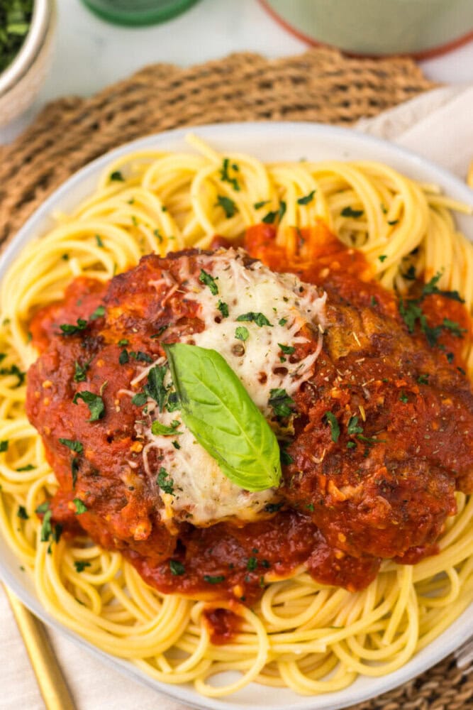 A plate of spaghetti topped with marinara sauce, a breaded meat patty, melted cheese, grated parmesan, chopped parsley, and a fresh basil leaf.