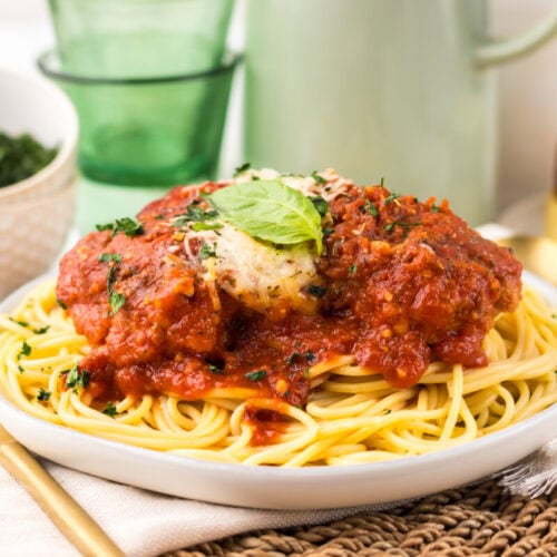 A plate of spaghetti topped with tomato sauce, meatballs, and a basil leaf, with a fork, knife, bread, and a bowl of greens in the background.