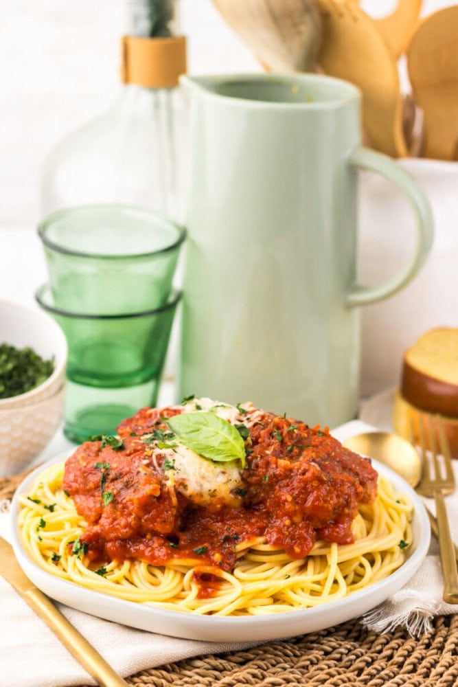 A plate of spaghetti topped with tomato sauce, chicken, and basil is set on a table with green glasses, a pitcher, and a small bowl of greens in the background.