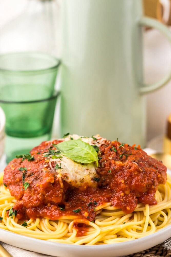 A plate of spaghetti topped with tomato sauce, grated cheese, and a fresh basil leaf, with green glasses and a jug in the background.