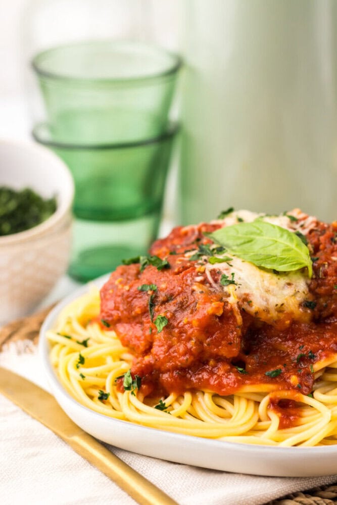 A plate of spaghetti topped with tomato sauce, a breaded chicken cutlet, grated cheese, and a fresh basil leaf, with glasses and a bowl in the background.