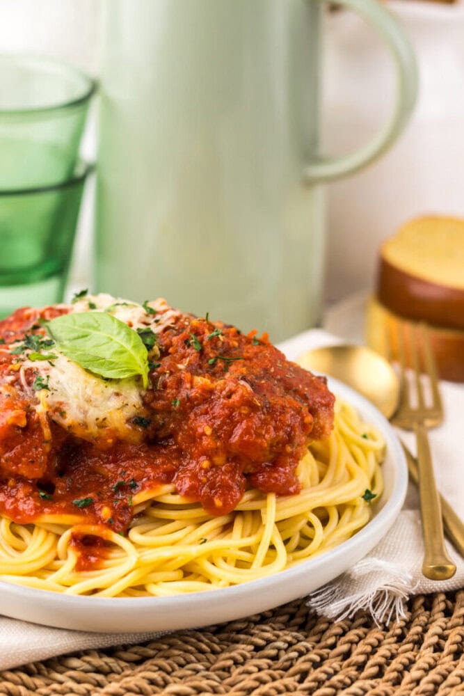 A plate of spaghetti topped with tomato sauce, herbs, and cheese, garnished with a basil leaf. A fork, bread, and a glass are in the background.