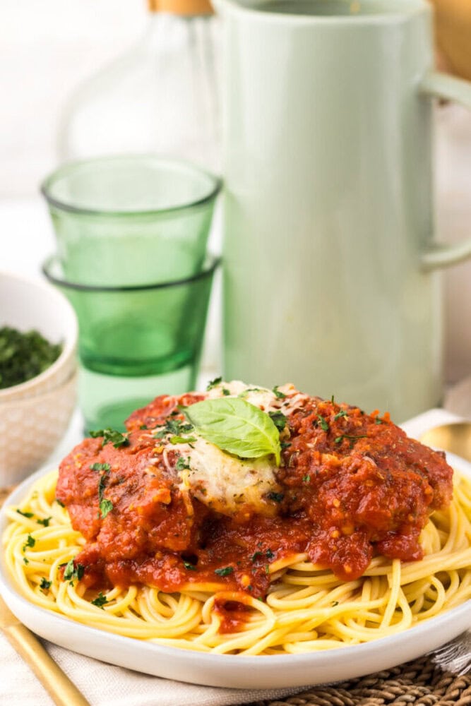 A plate of spaghetti topped with tomato sauce, melted cheese, and fresh basil, with green glasses and a pitcher in the background.