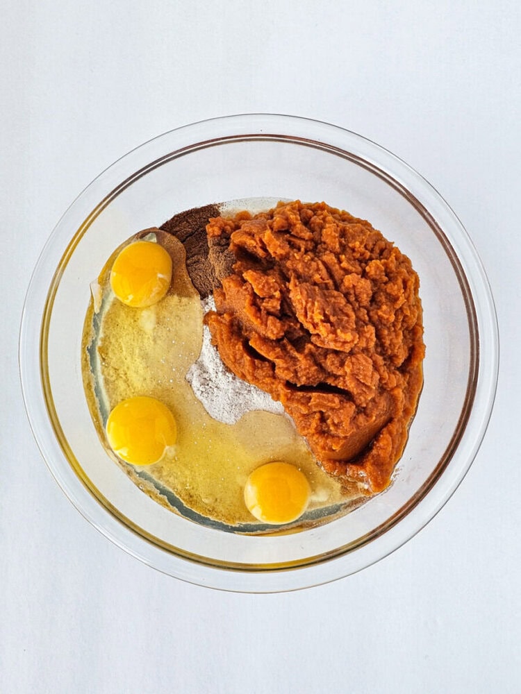 A glass bowl containing three eggs, pumpkin puree, and various dry ingredients on a white surface.