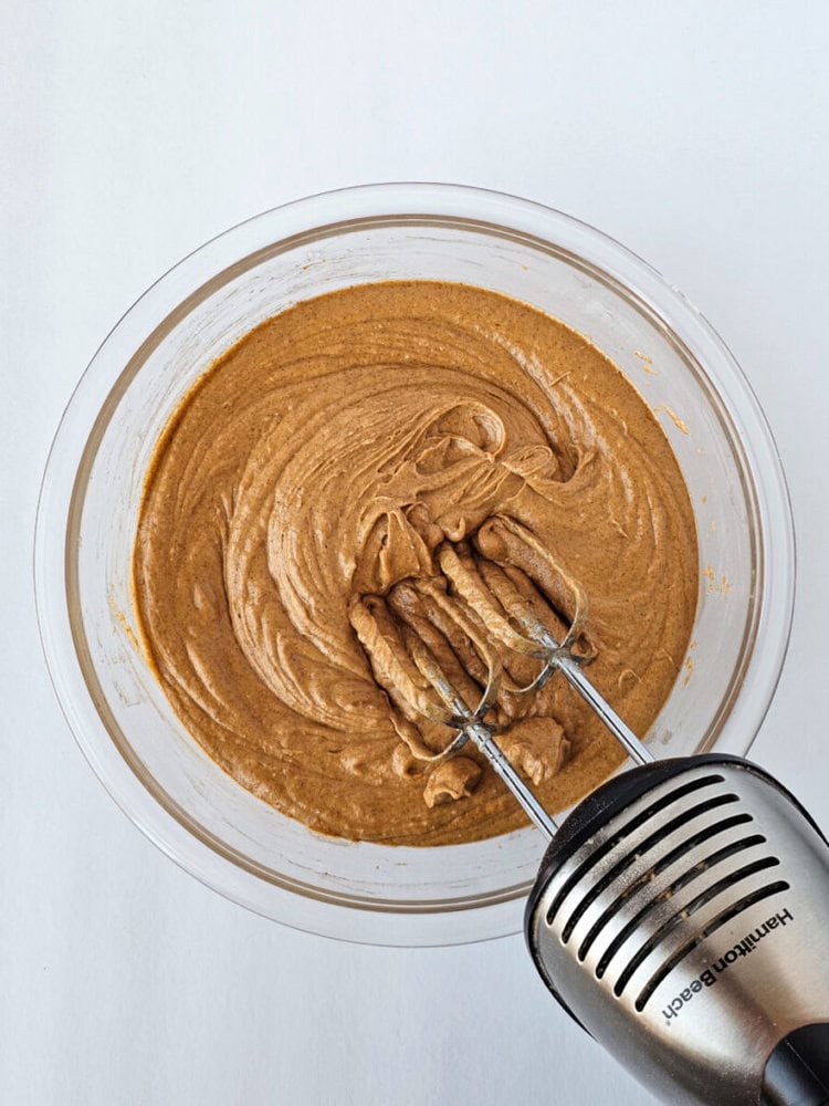 A hand mixer with beaters is immersed in a bowl of brown batter, which appears well-mixed, on a white surface.