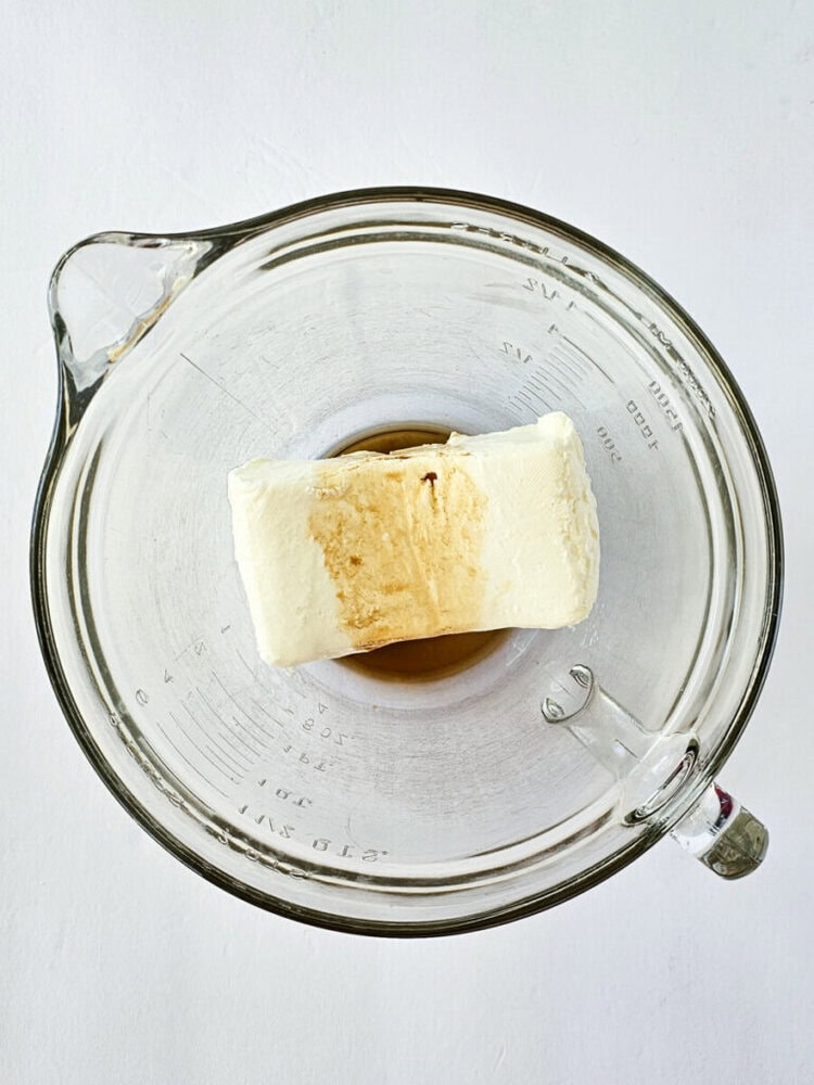 A block of cream cheese and vanilla extract in a glass measuring bowl, viewed from above, on a white surface.