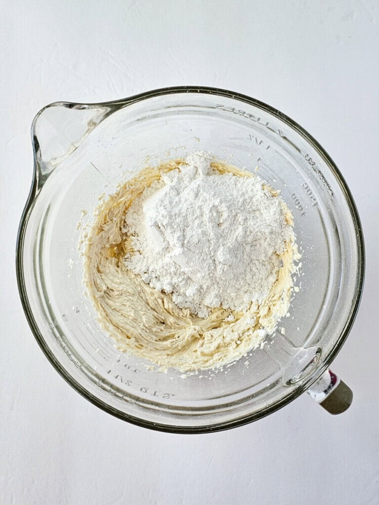 A glass mixing bowl containing creamed butter and sugar with a pile of flour on top, ready to be mixed together.