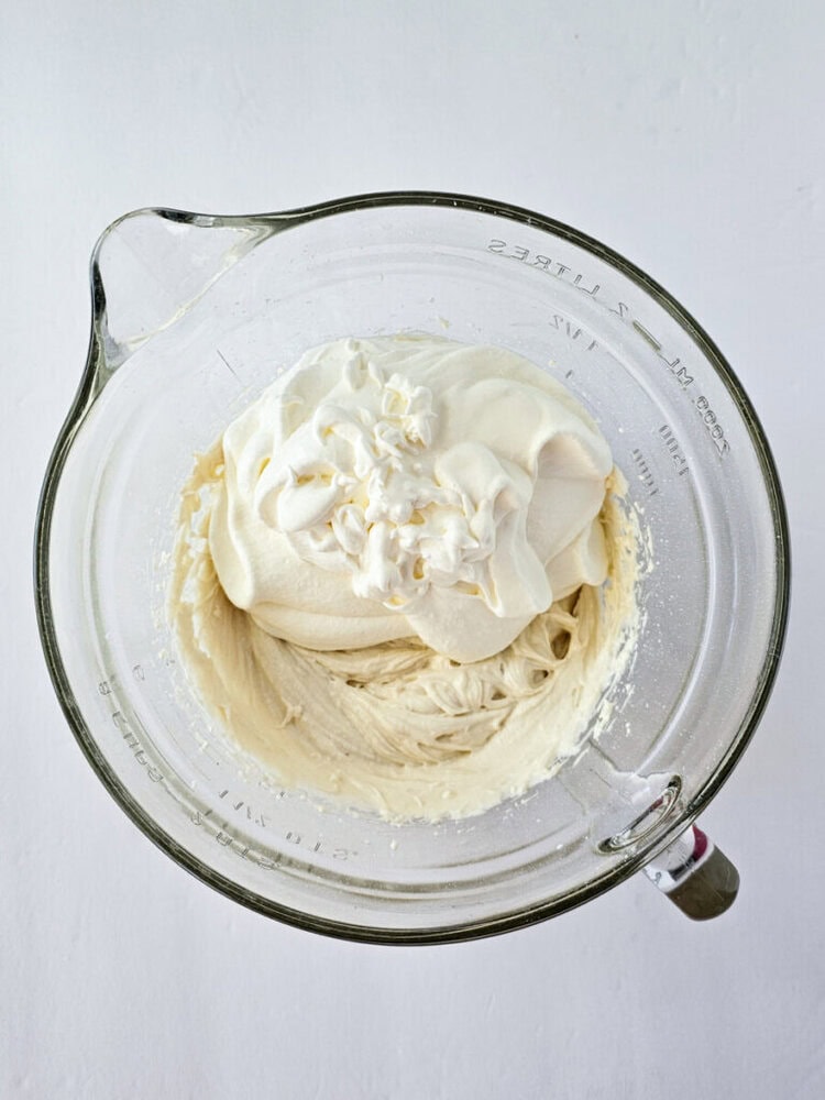 A glass mixing bowl filled with thick, whipped cream or frosting sits on a white surface.