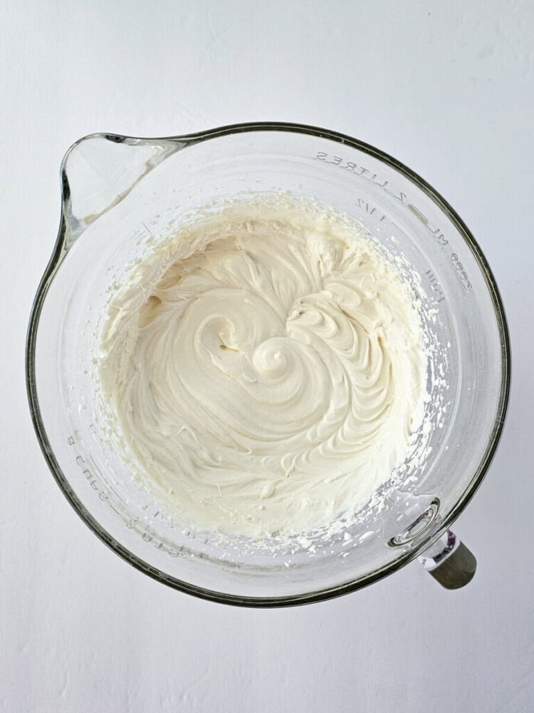 A glass mixing bowl filled with smooth, swirled, creamy white frosting on a white surface.