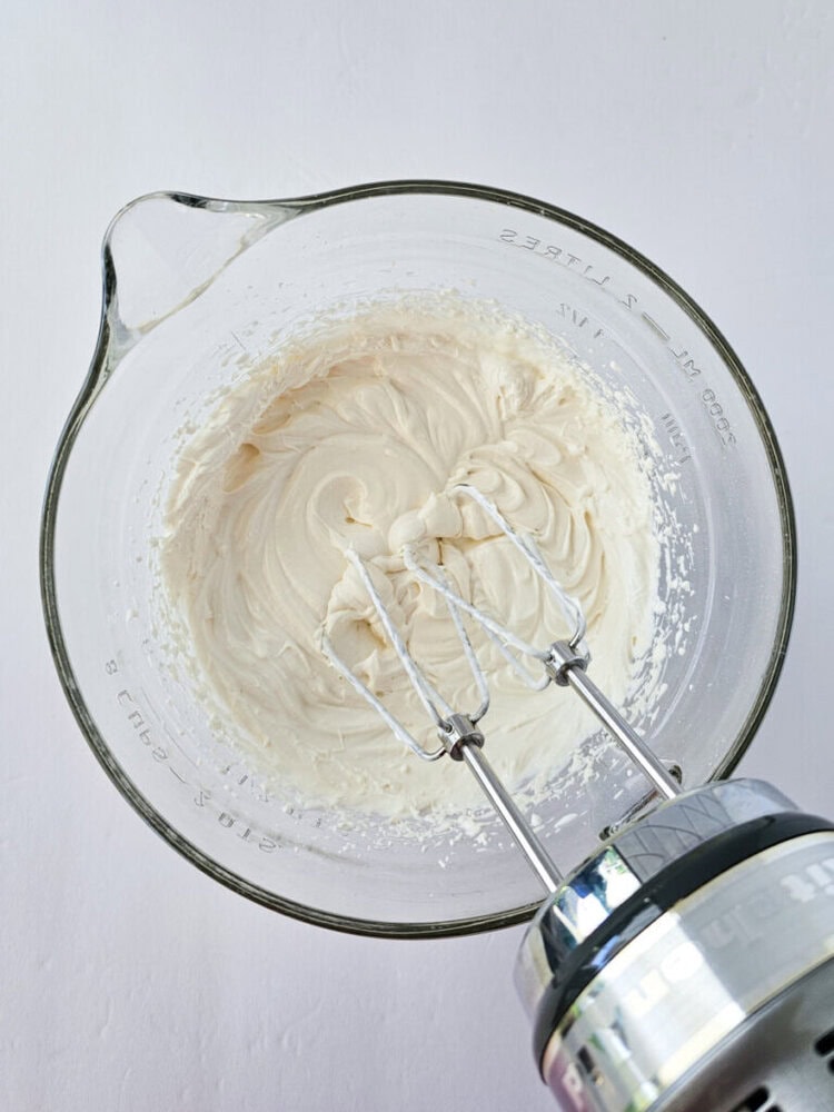 A glass mixing bowl filled with whipped cream cheese mixture, with electric mixer beaters resting inside.
