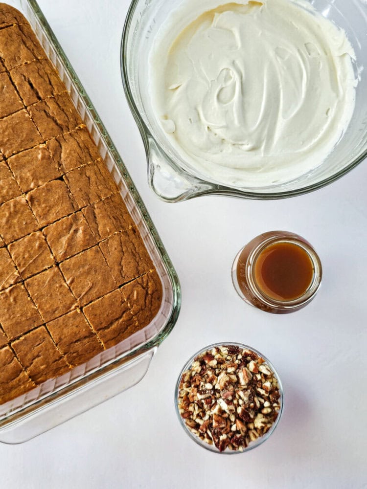 A glass baking dish with sliced cake, a bowl of whipped cream, a jar of caramel sauce, and a bowl of chopped pecans on a white surface.