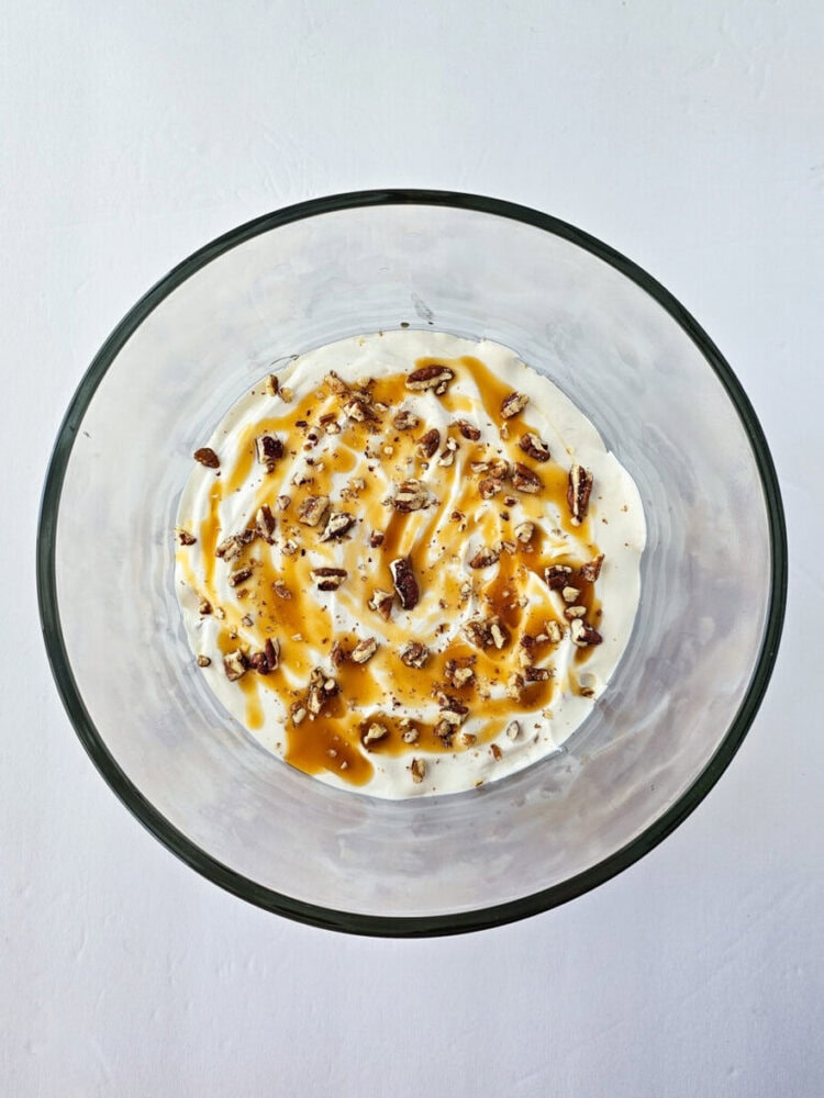 A glass bowl filled with creamy dessert topped with chopped nuts and a drizzle of caramel sauce, viewed from above.