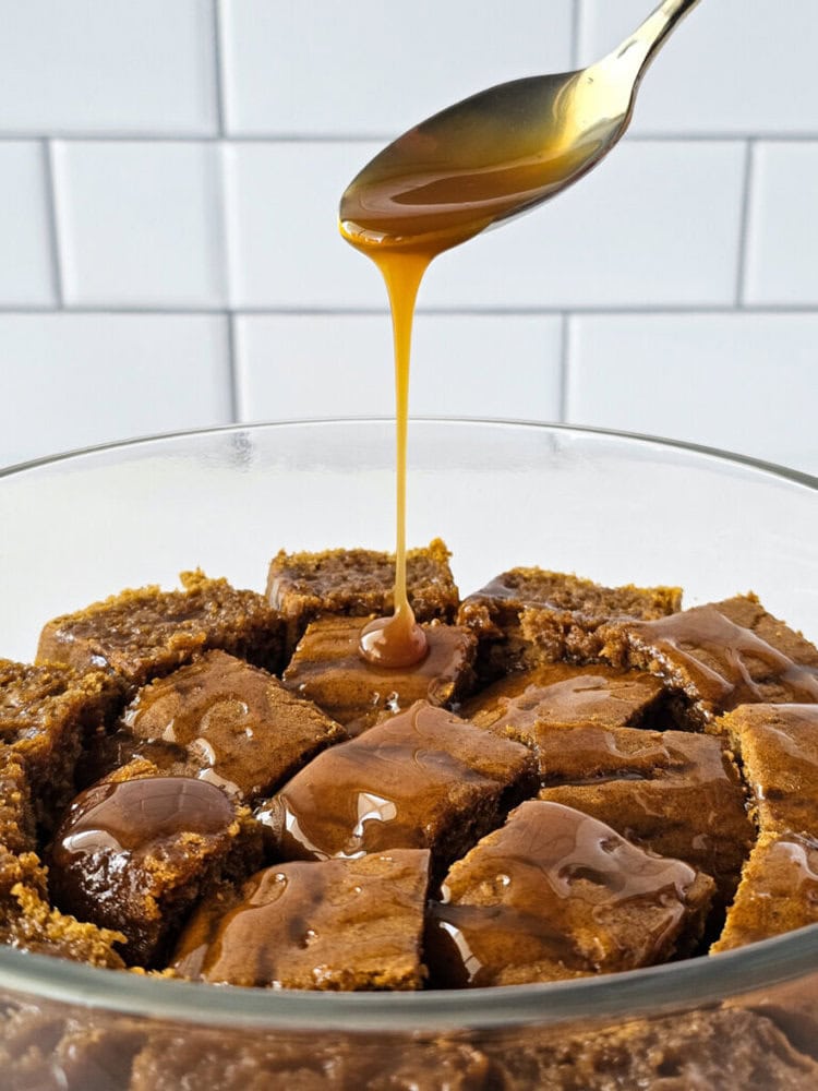 A spoon drizzles caramel sauce over a bowl of square-cut pieces of brown cake, with a white tiled background.