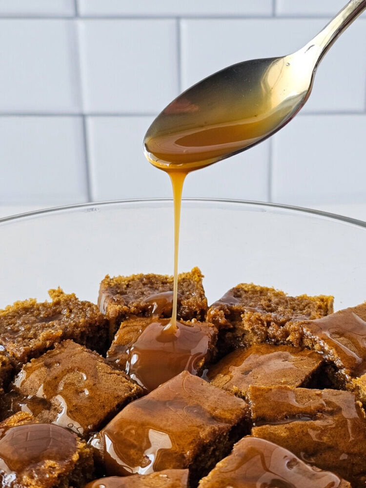 A spoon drizzles caramel sauce over a bowl of square-cut pieces of brown cake, with a white tiled wall in the background.