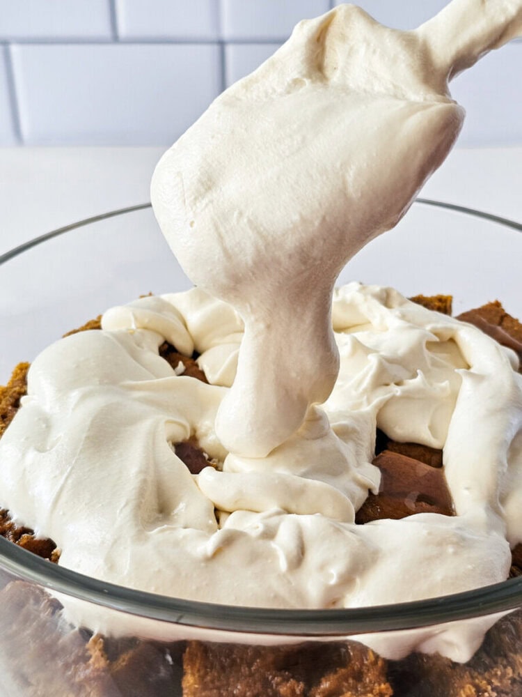 A spatula spreads thick, white cream over a layer of crumbled brown cake in a clear glass bowl.