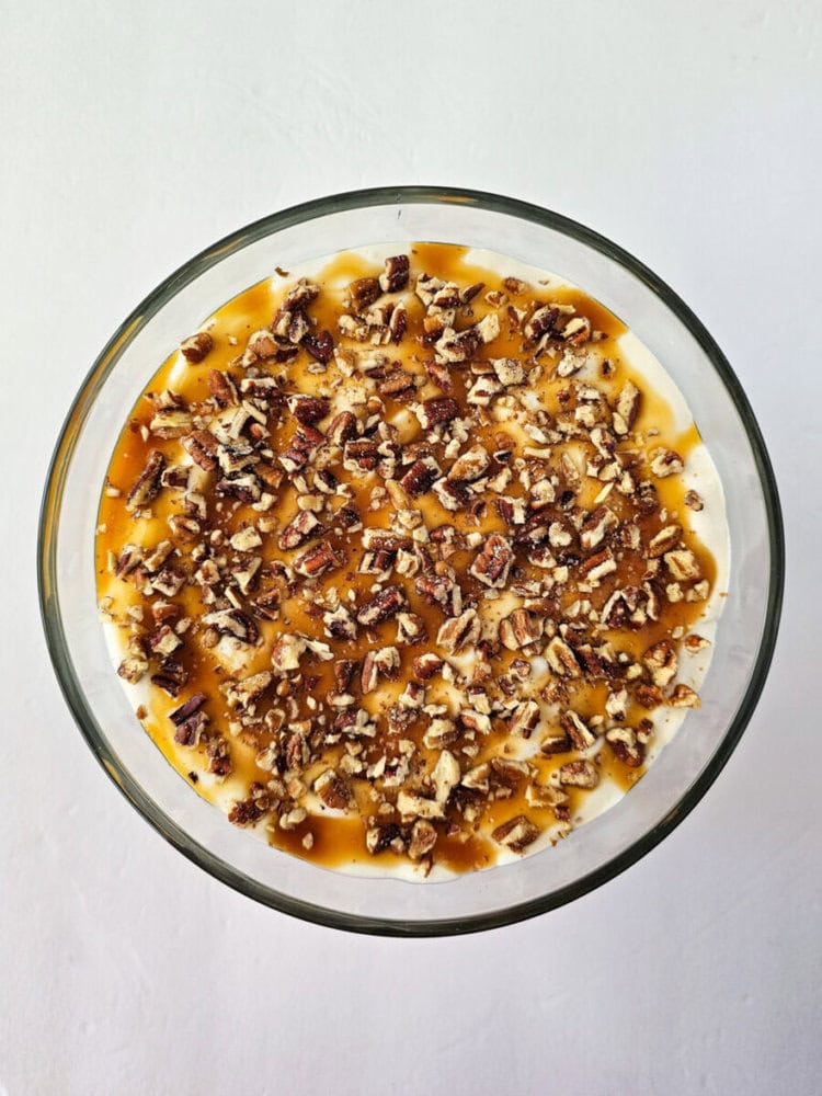 A glass bowl filled with creamy dessert, topped with chopped pecans and caramel sauce, on a white surface.