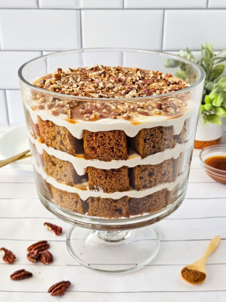 A trifle dessert in a glass bowl with layers of cubed cake, cream, and chopped pecans, set on a white surface with pecans and a spoon nearby.