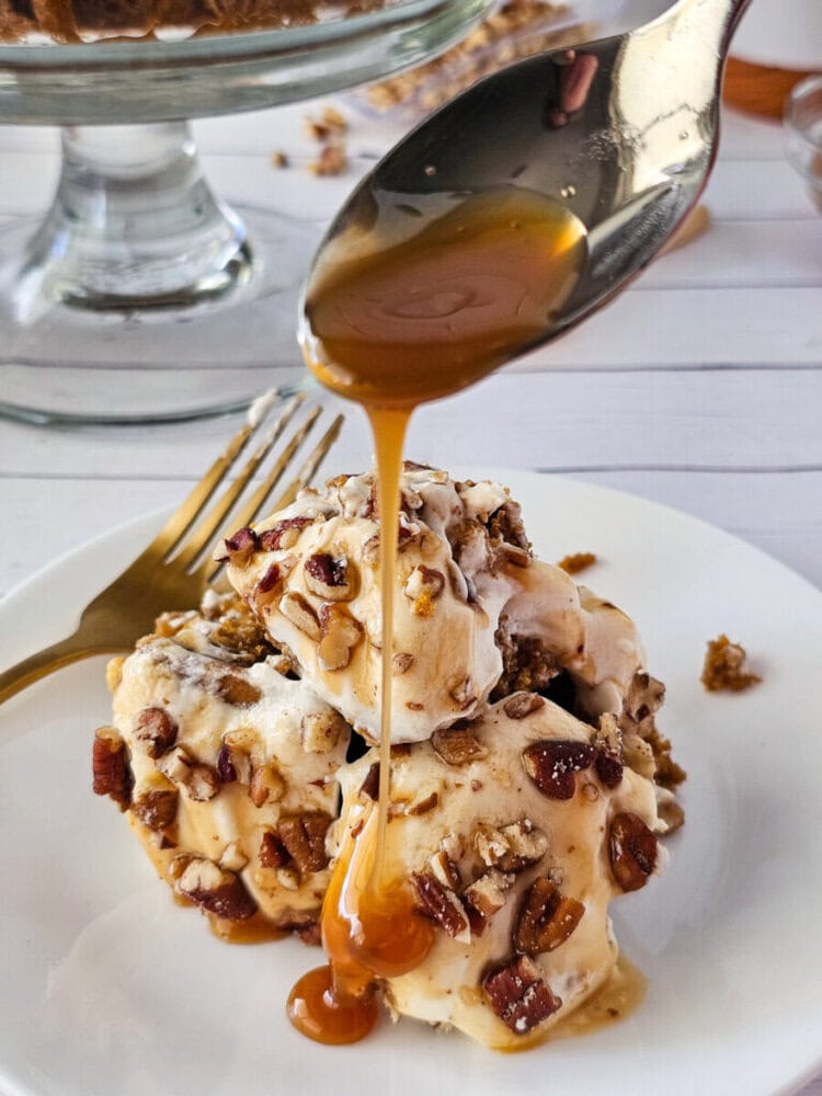 A spoon drizzles caramel sauce over a stack of ice cream topped with chopped nuts on a white plate, with a fork and dessert stand in the background.