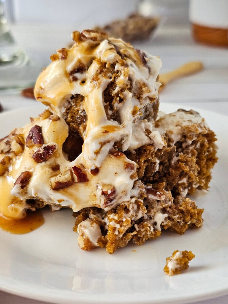 A close-up of a dessert featuring moist cake pieces topped with melting vanilla ice cream, pecans, and caramel sauce on a white plate.