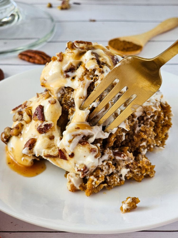 A fork cuts into a slice of spiced cake topped with creamy frosting, caramel sauce, and chopped pecans on a white plate.